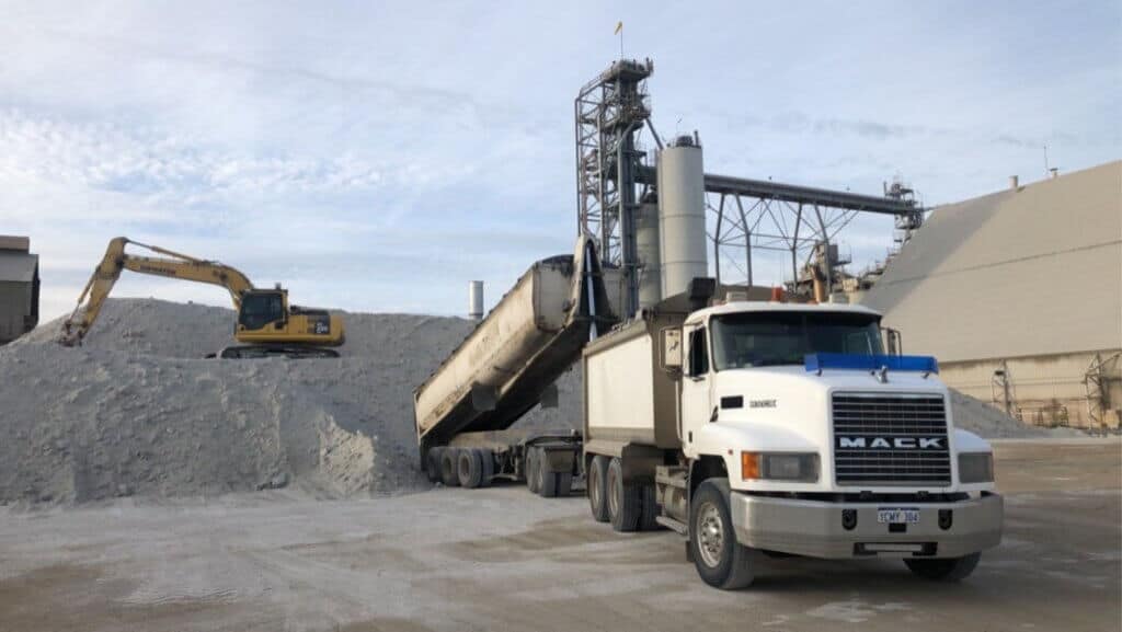 A large double dump body truck, the back dump body is in the process of offloading onto a large mountain of sand