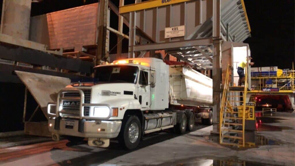 A large truck in an industrial yard, receiving goods into its storage holder